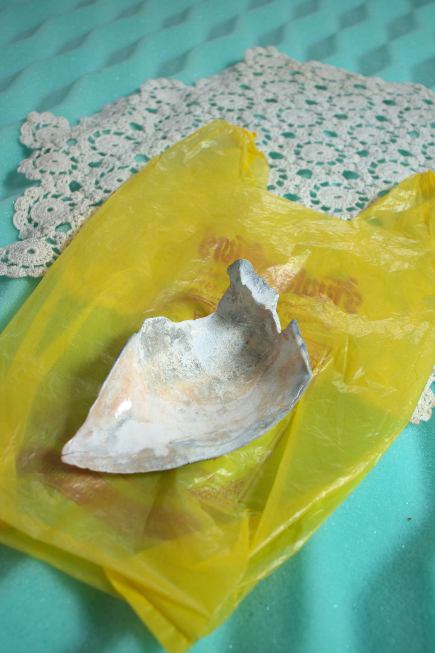 Seashell fragment laying on top of yellow plastic bag over a doily on a bright blue background
