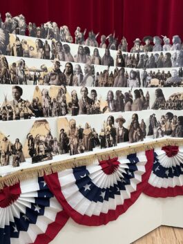 Cut out black and white portraits set in a riser-like formation in front of heavy red drapes with half-circle red, white, and blue banners below