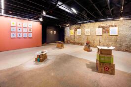 Raw gallery space with coral-colored wall in back left and exposed brick on the right displaying framed objects and sculptures on a concrete floor.